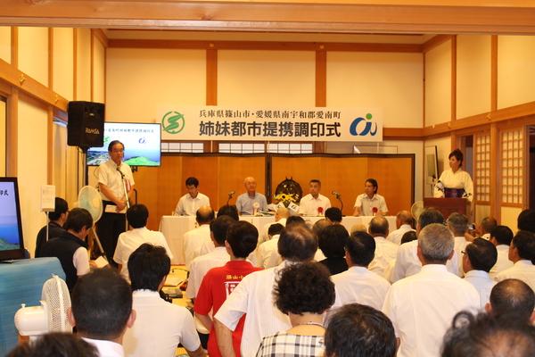 姉妹都市提携調印式で男性がスタンドマイクで話し、それを静かにお客さんが座って聞いている写真