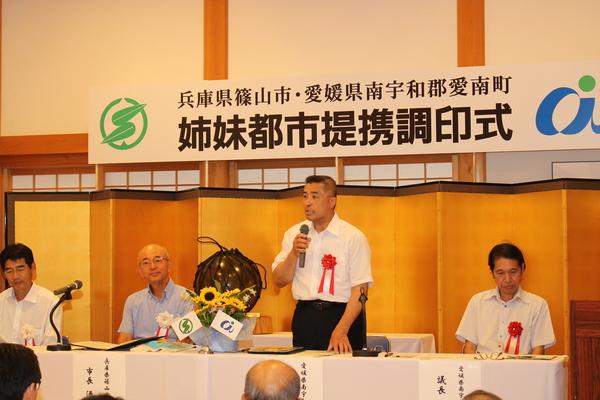 姉妹都市提携調印式で男性がマイクを手に持ち、お話しして同じテーブルに座っている方々は聞いている写真