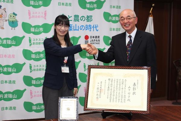 賞状と記念盾が飾られている後ろで市長と山下さんが握手をして記念撮影をしている写真