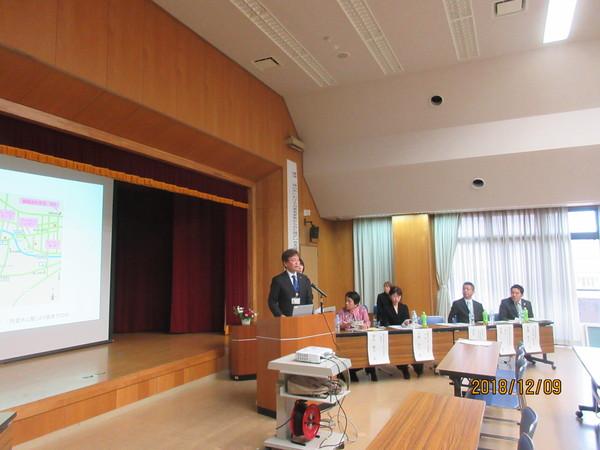 左にモニターがあり、その右にある演台で男性が立ちながらマイクで話をしている写真