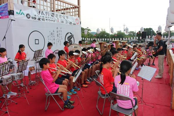 やぐらの下でピンクや赤のTシャツをきた小学生が吹奏楽の演奏をしている様子を横から写した写真