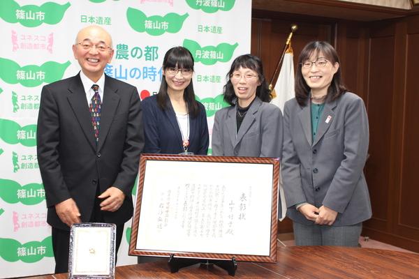 篠山市と書かれた壁の前で、賞状と一緒に市長、山下さん、スーツを来た女性2人が微笑んで記念撮影をしている写真