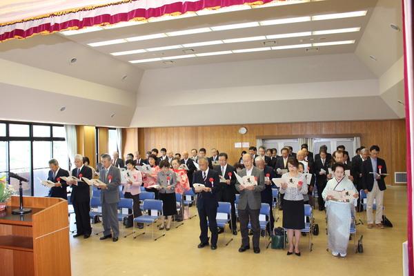 胸にリボンを付けた受賞者全員が席を立ち冊子を読み上げている写真