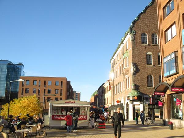 青空の下、茶色の建物の横を上下黒の服をきた男性が歩いており飲食コーナーの椅子に座っている人達小さなお店の前で並んでまっている写真