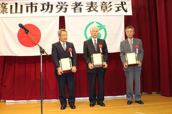 壇上で受賞者男性3名が賞状の入った盾を持っている写真