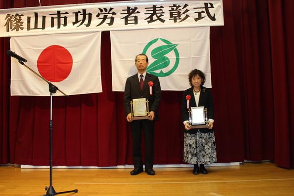壇上で受賞者男性1名、女性1名が賞状の入った盾を持っている写真