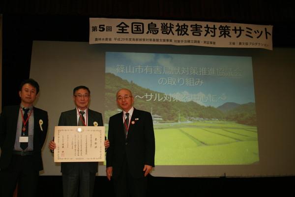 第5回 全国鳥獣被害対策サミットと書かれている横断幕の前で、西潟さんが表彰状を手に持って市長と鈴木 克哉さんと写っている写真
