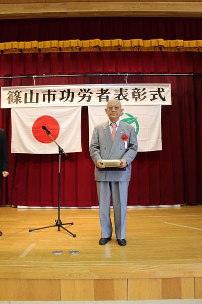 壇上で眼鏡をかけ、グレーのスーツを着た受賞者男性1名が賞状の入った盾を持っている写真