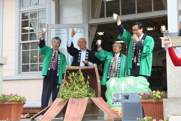 中央に枝豆の入った樽があり、その後ろで4人の法被を着た男性達が片手で紙コップを掲げている写真