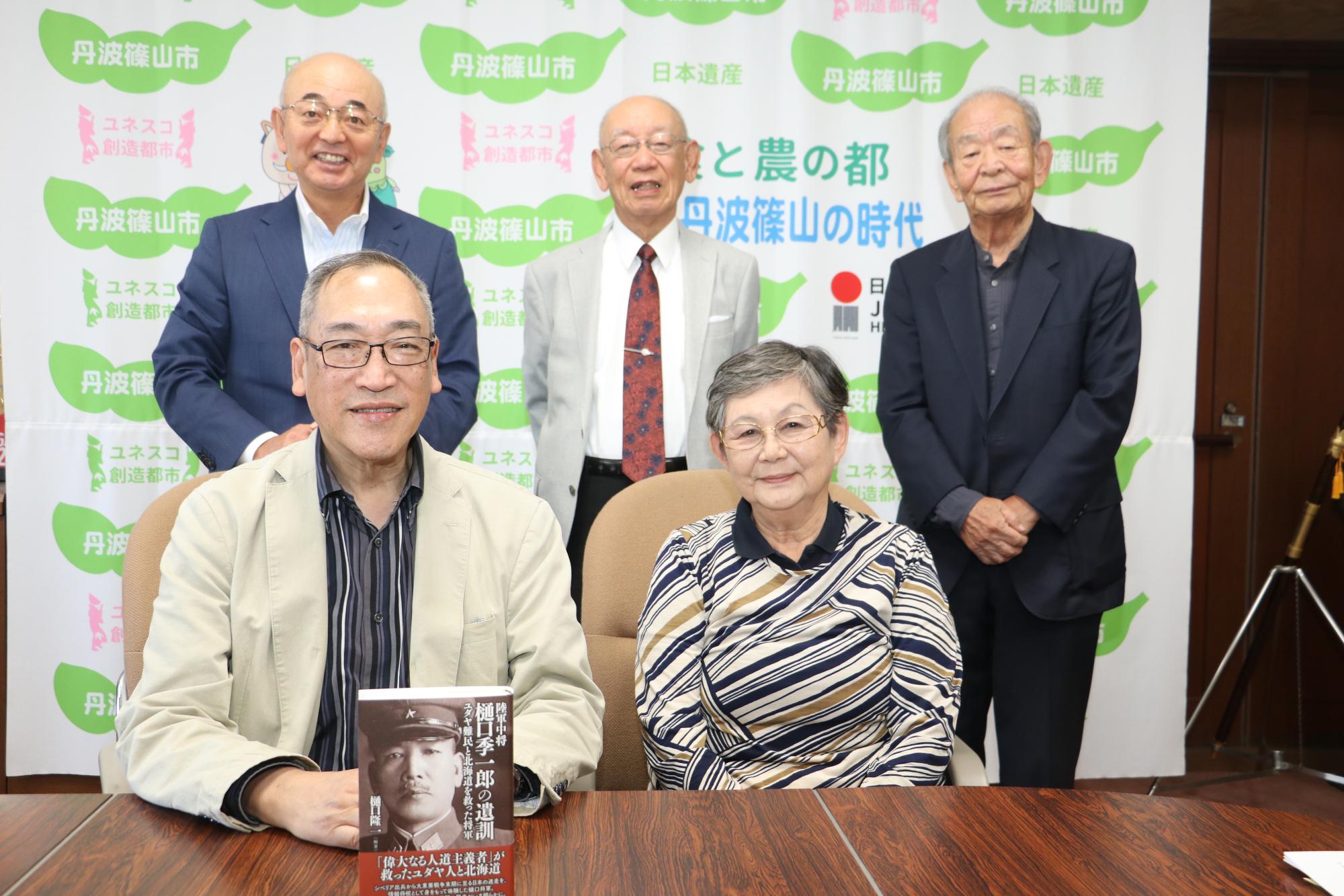 樋口夫妻が椅子に座られて、著書を手に持たれている。後方に、市長、小山さん、小林さんが並んで立っている。