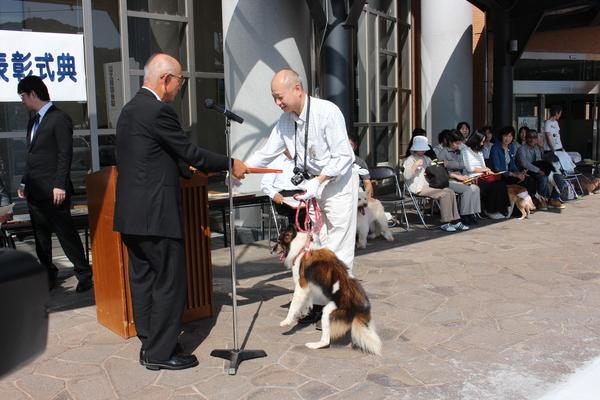 首にカメラを提げて手には首輪をした犬のリードを持っている飼い主さんへ市長が賞状を手渡している写真