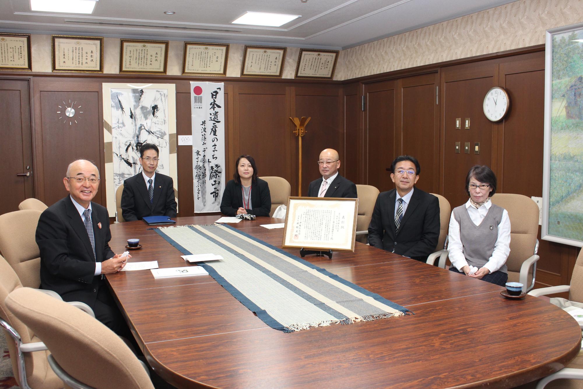 円卓に座って市長と国際理解センター、市民協働課の職員が記念撮影、机上には表彰状が飾ってある