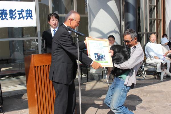 黒い色の長寿犬を膝を曲げながら抱っこした飼い主さんへ額に入った賞状を手渡している市長の写真