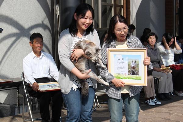 額に入った賞状を持った女性と長寿犬を抱っこしている女性の写真