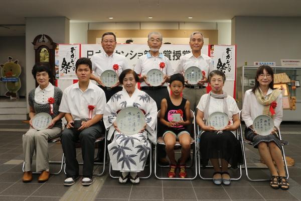 歌詞の書かれた皿やコーヒーカップを持った9名の受賞者たちの集合写真