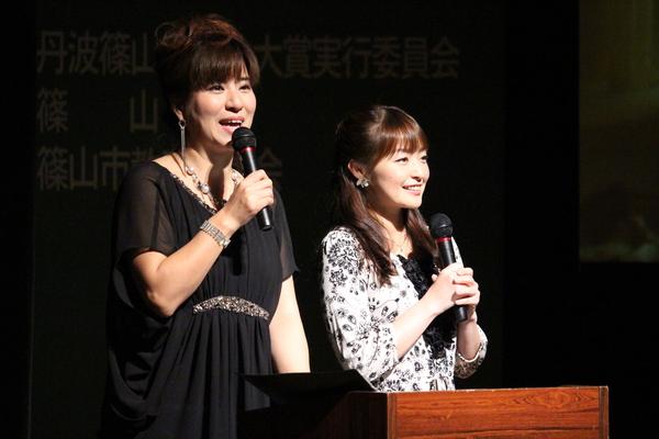 演台の前で、黒いワンピースを着た女性がマイクを持って話をしており、その横に白地に ペイズリー柄のワンピースを着た女性がマイクを持っている写真