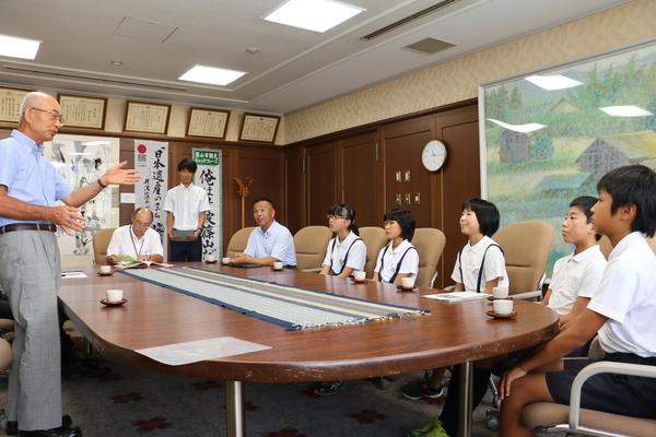 応接室の、会議テーブルに座る小学生の生徒たちと、関係者、その前で立って話をしている市長の写真