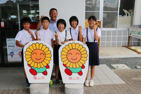 花のイラストが描かれた看板の前で、ガッツポーズをする小学生の生徒と、男性の写真