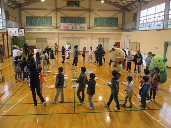 小学生やまるいのの着ぐるみたちが体育館で円になって歩いている写真