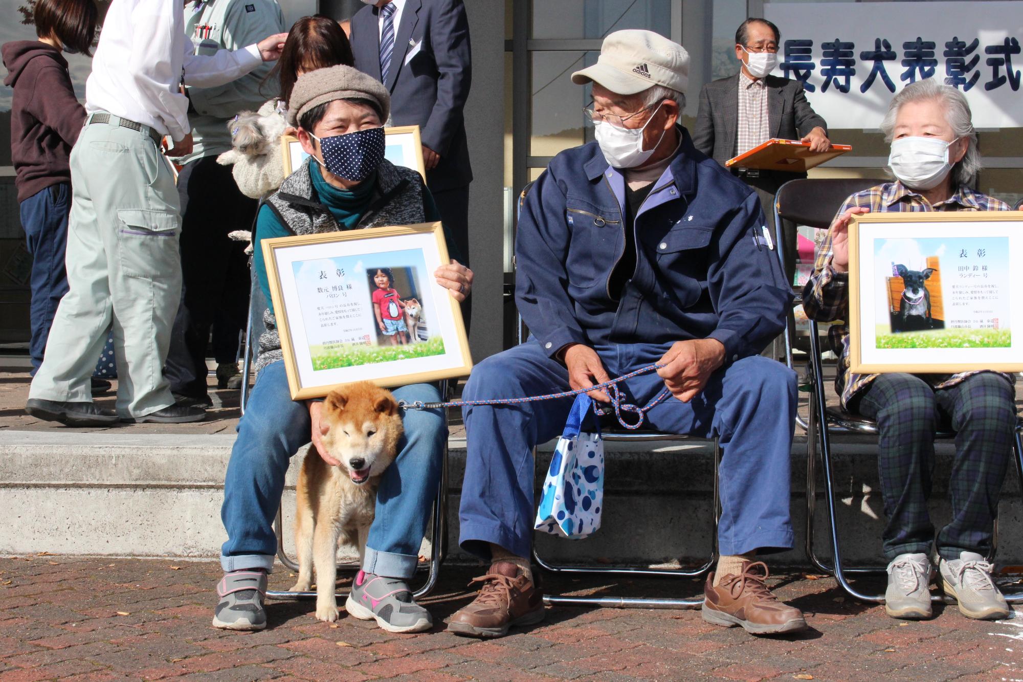 椅子に座って、飼い犬をなでながら受け取った表彰状を見る飼い主たち