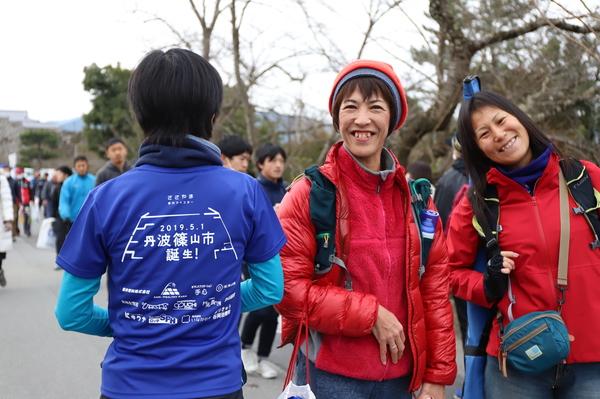 丹波篠山誕生のTシャツを着た後ろ姿の人の横で赤色の上着を着た2名の女性が笑顔で写っている写真