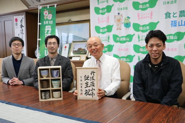 丹波篠山市誕生祝と書かれた木箱の蓋を持った市長と、男性3人が座っている写真