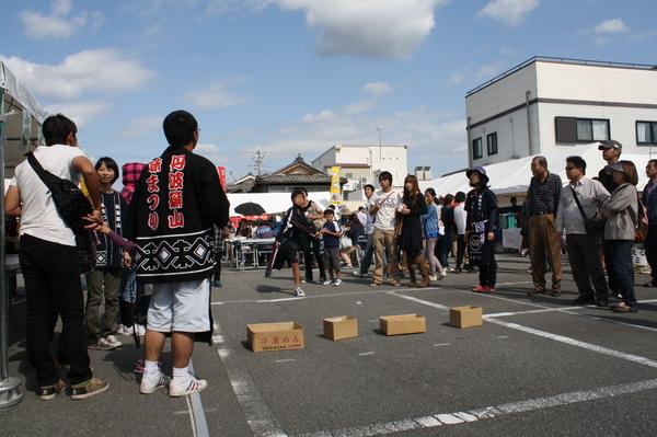 さや投げコンテストで男の子が設置されている段ボールをめがけてさやを投げ、それを見ている観客の人達の写真