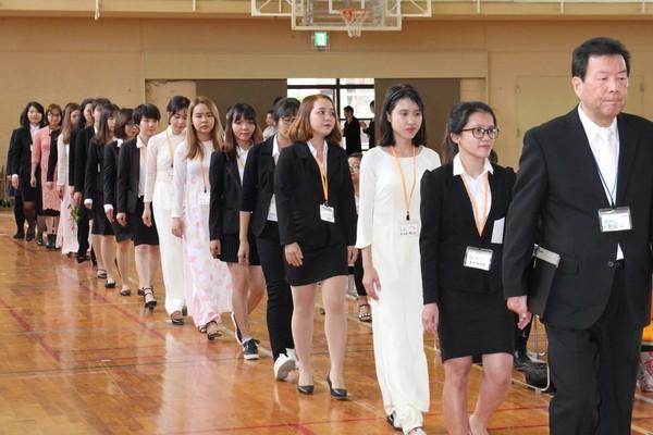 体育館の中をスーツや白いドレスを着た女性たちが縦一列に並んで歩いている写真