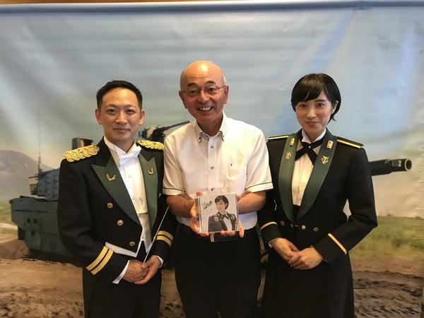 戦車の写真の前で、市長が鶫（つぐみ）さんのCDを手に持ち黒い礼服を着ている柴田さん、鶫（つぐみ）さんと一緒に写っている写真
