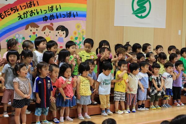 ともだちいっぱい、えがおいっぱい、ゆめいっぱいの文字と子供の絵が描かれた壁紙の前に40名ほどの園児が横3列に並んでいる写真