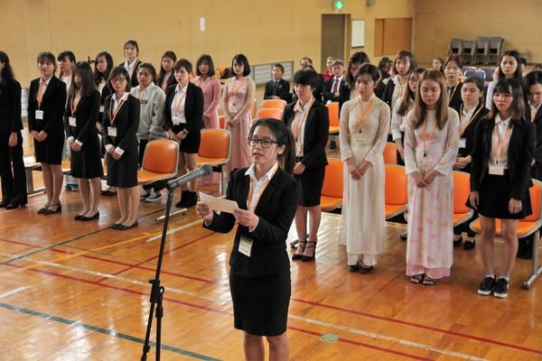 後ろに女子学生たちが立っていて、代表の女性が紙を持ちながらマイクで話をしている写真