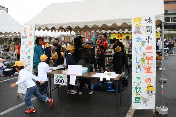 白いテントの横に味間小 三年生のおいしい黒枝豆と書かれたのぼり棒が設置されており、黄色い帽子を被った小学生と大人の人が集まっている写真