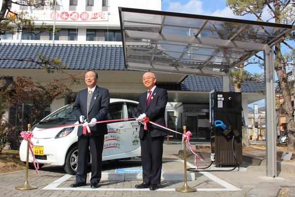 白い軽の公用車が停まっている急速充電器の前で市長と関係者の男性が紅白リボンを手に持ってテープカットをしようとしている様子の写真