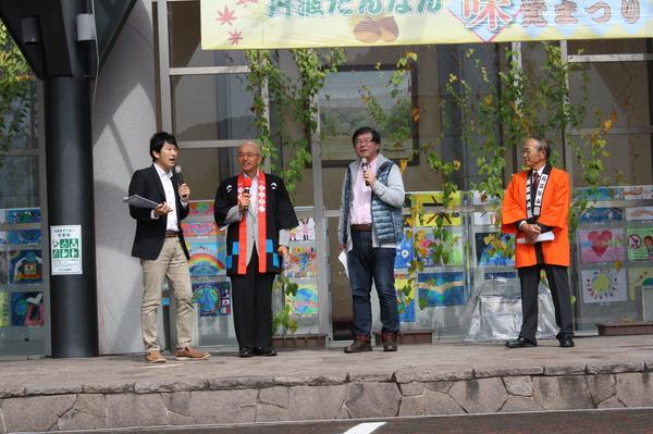 石段の上に市長と市長の両脇にマイクを持って話をしている男性2人がおり、オレンジの法被を着て手を前に組んでいる男性が写っている写真