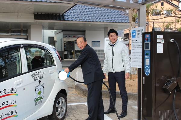 白い軽の公用車にスーツを着た男性が急速充電器を使って充電しており、白いジャンパーを着た男性が充電している様子を見守っている写真