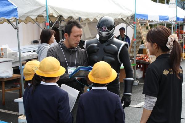 黄色の帽子を被った3名の小学生の横に紙を結んだ女性が立っており、篠山市の戦士 ササグローと男性が話している様子を見ている写真