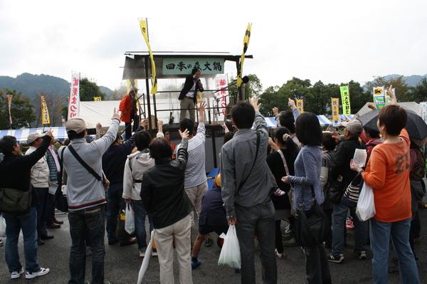 木の舞台上に上がっている男性が集まった観客の方とじゃんけんをしている様子の写真