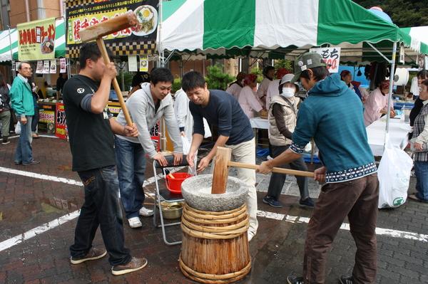 2人の男性が、みかげ石の臼で杵を打ちながら交互に餅をつき、餅に水をつけている男性と椅子に両手を置いてその様子を見ている男性の写真