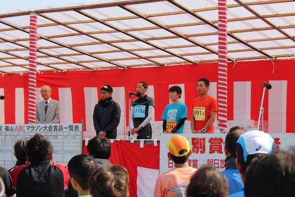 紅白の幕が張られた舞台の上に市長と4名の選手が立っていて、その中の一人の選手がマイクで話している写真