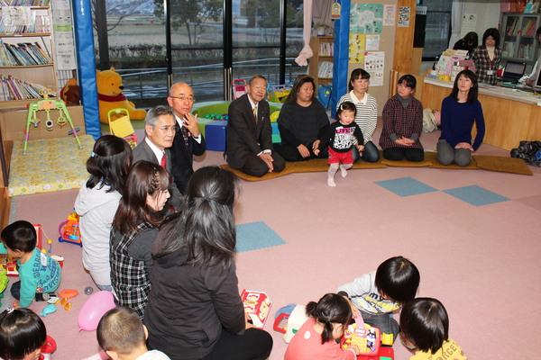 輪になって座って話し合いをしている母親たちと市長と議員の方と、そばで遊んでいる子供たちの写真