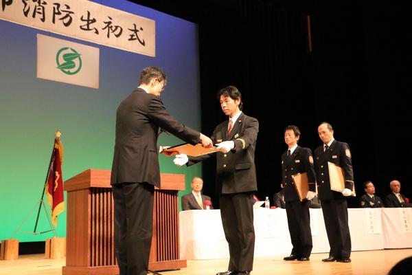 出初式にて壇上で制服姿の男性が表彰されている写真