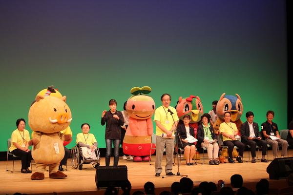 ステージの上で黄色いTシャツを着た男性がマイクで話していて、その横で手話で説明する女性や着ぐるみが立っている写真