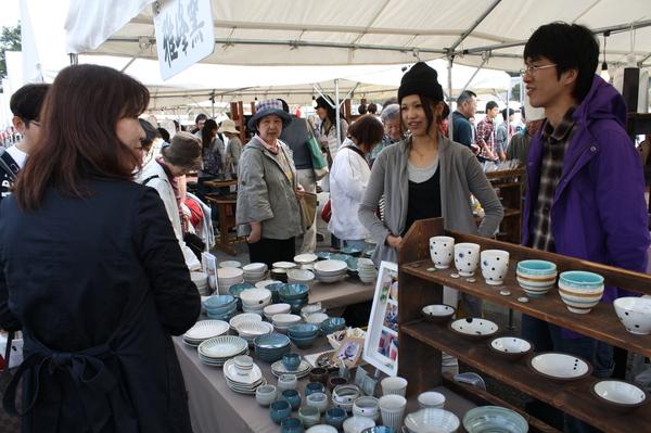 机の上に並べられた、陶器のお皿やカップを見ているお客さんや、店員と話す女性の写真