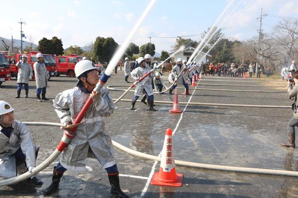 消防団員らが消防車のホースを持ち、放水演習を行っているのを右側から撮影されている写真