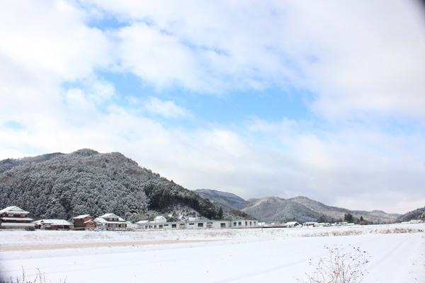空の雲の合間から青空が見えており、山々や家々には雪が積もっており、田んぼにも真っ白な雪が降り積もっている様子の写真