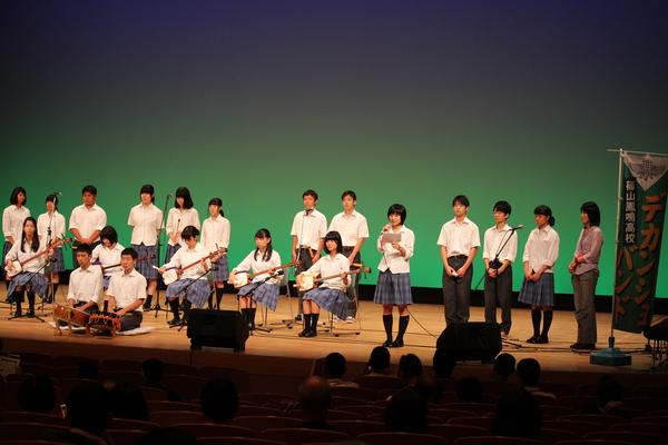 ステージの上で学生が20名ほど並んでいて、5名が三味線、2名が太鼓、その他の人たちが笛を持っている写真
