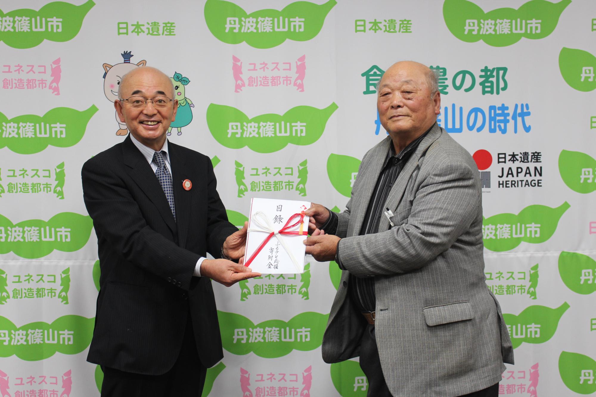 井関さんが寄付目録を市長に手渡そうとしている。