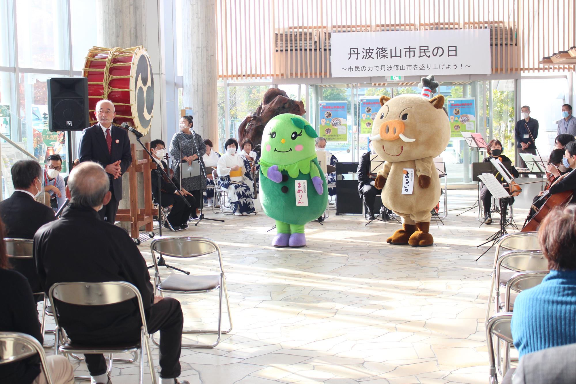 玄関ホールでの式典、パイプ椅子に座っている市民の方の前であいさつする市長、そばにはまるいのとまめりん。