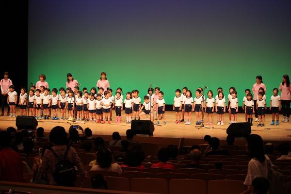ステージの上で30名ほどの白い体育着を来た幼稚園生や、ピンクのポロシャツを来た先生が並んでいる写真