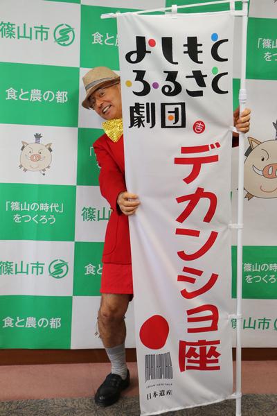 赤い衣装に黄色い蝶ネクタイをし帽子を被った市長がデカンショ座ののぼり旗の後ろから顔と体右半分を出している写真